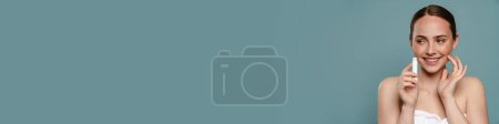 Téléchargez les photos : Jeune femme aux cheveux bruns souriants montrant un bâton de baume à lèvres debout sur fond bleu - en image libre de droit