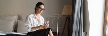 Téléchargez les photos : Femme d'affaires blanche brune d'âge moyen assise sur le lit dans une chambre d'hôtel tenant un téléphone portable - en image libre de droit