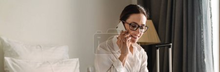 Foto de Sonriente de mediana edad morena blanca mujer de negocios sentada en la cama en una habitación de hotel hablando en el teléfono móvil utilizando el ordenador portátil - Imagen libre de derechos