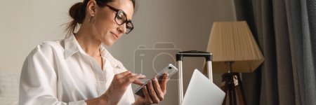 Téléchargez les photos : Femme d'affaires blanche brune d'âge moyen assise sur le lit dans une chambre d'hôtel tapant sur un téléphone mobile travaillant sur un ordinateur portable - en image libre de droit