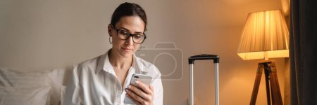 Photo for Mid aged brunette white businesswoman sitting on bed in a hotel room typing on mobile phone working on laptop - Royalty Free Image