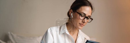 Foto de Mediados de edad morena blanca empresaria sentada en la cama en una habitación de hotel tocando tableta dispositivo con auriculares inalámbricos - Imagen libre de derechos