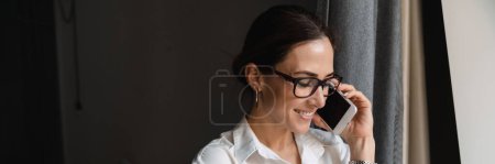 Photo for Smiling middle aged brunette white businesswoman in smart wear standing at the window in the hotel room talking on mobile phone - Royalty Free Image