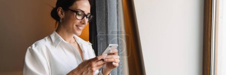 Téléchargez les photos : Brune d'âge moyen souriante femme d'affaires blanche en tenue intelligente debout à la fenêtre dans la chambre d'hôtel tenant un téléphone portable - en image libre de droit