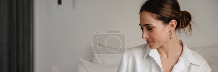 Photo for Mid aged brunette white businesswoman sitting on bed in a hotel room typing on laptop holding diary - Royalty Free Image