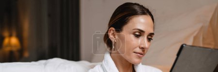 Photo for Smiling middle aged brunette white woman in bathrobe sitting on a floor at the bed indoors holding tablet computer - Royalty Free Image