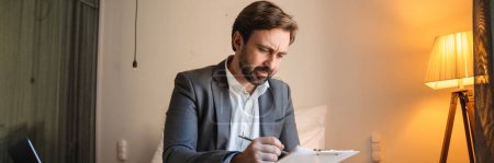 Photo for Mid aged bearded man in formal wear sitting on bed with laptop computer in the hotel room taking notes - Royalty Free Image