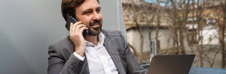 Foto de Sonriente hombre de negocios de mediana edad sentado en la terraza del hotel hablando por teléfono móvil - Imagen libre de derechos