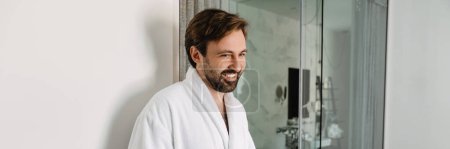 Téléchargez les photos : Homme blanc barbu souriant en peignoir prenant une tasse de café le matin dans la chambre d'hôtel - en image libre de droit