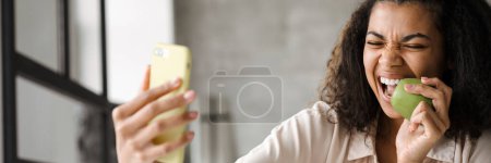 Foto de Joven mujer afroamericana feliz sosteniendo el teléfono inteligente haciendo un vlog desde casa mordiendo manzana verde - Imagen libre de derechos