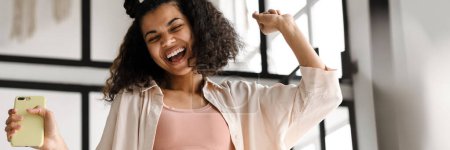 Téléchargez les photos : Jeune femme heureuse afro-américaine tenant téléphone intelligent dansant en s'amusant à la maison - en image libre de droit