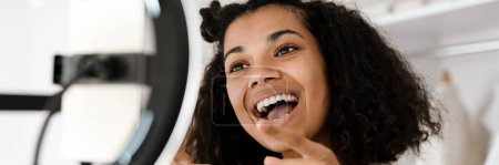 Téléchargez les photos : Heureuse jeune femme africaine influenceuse debout devant une caméra à la maison faisant vlog - en image libre de droit