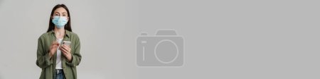 Téléchargez les photos : Positif jeune femme blanche portant un masque médical de protection hoding téléphone portable debout sur le mur gris - en image libre de droit