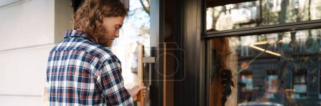 Photo for Young white cafe worker in plaid shirt opening door outdoors - Royalty Free Image