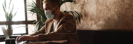Téléchargez les photos : Jeune homme blanc dans le masque de visage travaillant avec ordinateur portable dans le café - en image libre de droit