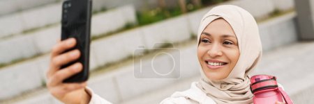 Téléchargez les photos : Moyen-Orient femme prendre selfie sur téléphone mobile tout en travaillant à l'extérieur - en image libre de droit