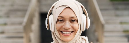 Foto de Mujer de Oriente Medio en hijab escuchando música con auriculares inalámbricos al aire libre - Imagen libre de derechos