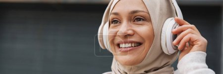 Foto de Mujer de Oriente Medio en hijab riendo mientras escucha música con auriculares inalámbricos al aire libre - Imagen libre de derechos