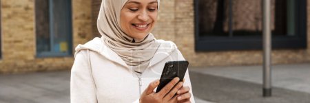 Foto de Mujer de Oriente Medio en hijab usando teléfono móvil y sonriendo al aire libre - Imagen libre de derechos