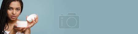 Téléchargez les photos : Jeune femme noire avec des nattes montrant crème pour le corps isolé sur fond bleu - en image libre de droit