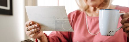 Téléchargez les photos : Vue rapprochée d'une femme âgée regardant de vieilles photos avec une tasse dans les mains alors qu'elle était assise dans un café - en image libre de droit