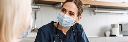 Téléchargez les photos : Une femme médecin souriante dans un masque protecteur jetable à l'écoute des plaintes de sa patiente âgée à la maison - en image libre de droit