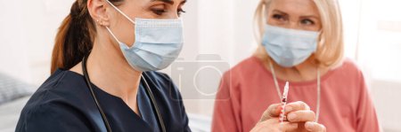 Photo for A female doctor in a protective disposable mask wants to give insulin injection to her elderly patient in the light room - Royalty Free Image