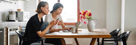 Téléchargez les photos : Une vue latérale du couple lesbien concentré regardant l'ordinateur portable et le téléphone assis dans la cuisine légère - en image libre de droit