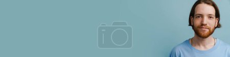 Téléchargez les photos : Jeune homme roux avec barbe posant et regardant la caméra isolée sur fond bleu - en image libre de droit