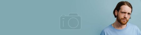 Téléchargez les photos : Jeune homme roux avec barbe fronçant les sourcils et regardant la caméra isolée sur fond bleu - en image libre de droit