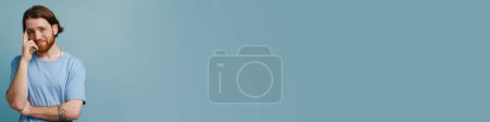 Téléchargez les photos : Jeune homme roux avec barbe souriante et regardant la caméra isolée sur fond bleu - en image libre de droit