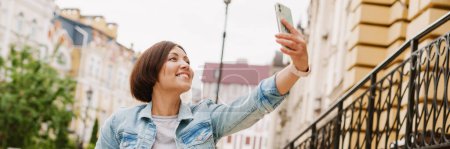 Téléchargez les photos : Brunette femme prendre selfie sur téléphone portable tout en étant assis en fauteuil roulant à la rue de la ville - en image libre de droit