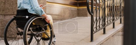 Téléchargez les photos : Brunette femme utilisant un téléphone portable assis en fauteuil roulant dans la rue de la ville - en image libre de droit