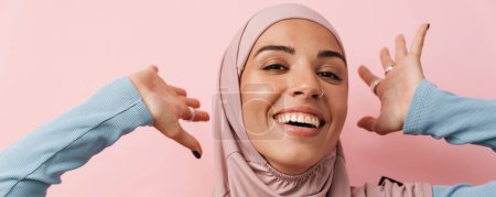 Foto de Un retrato de la mujer musulmana riéndose vestida con hiyab rosa de pie con los brazos levantados en el estudio rosa - Imagen libre de derechos