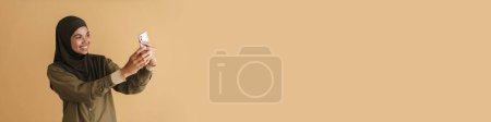 Téléchargez les photos : Femme musulmane noire en hijab prenant selfie sur téléphone mobile isolé sur fond beige - en image libre de droit