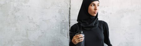 Téléchargez les photos : Jeune femme musulmane en hijab debout avec tapis de fitness et bouteille d'eau à l'intérieur - en image libre de droit