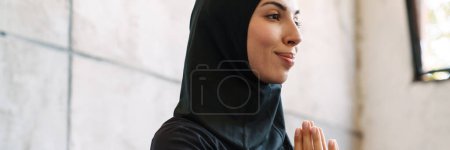 Téléchargez les photos : Jeune femme musulmane dans le hijab méditant tout en étant assis à l'intérieur - en image libre de droit