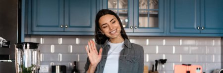 Photo for Smiling woman gesturing while taking selfie footage on cellphone at home kitchen - Royalty Free Image