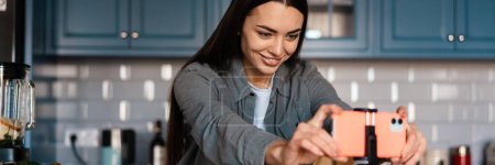 Téléchargez les photos : Femme souriante prenant des images selfie sur son téléphone portable tout en faisant du smoothie à la maison cuisine - en image libre de droit