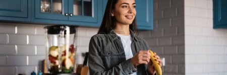 Téléchargez les photos : Femme brune blanche souriante tout en faisant un smoothie à la maison cuisine - en image libre de droit