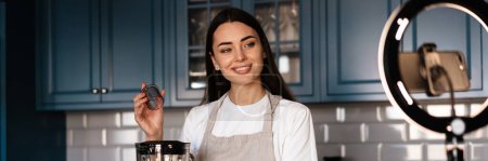 Foto de Mujer sonriente haciendo batido mientras toma imágenes de selfies en el teléfono celular en la cocina casera - Imagen libre de derechos