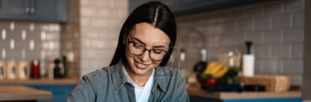 Photo for White smiling woman writing down notes while working with laptop at home kitchen - Royalty Free Image