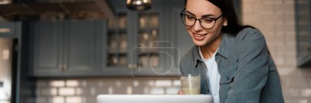 Foto de Mujer sonriente blanca bebiendo batido mientras trabaja con el ordenador portátil en la cocina del hogar - Imagen libre de derechos