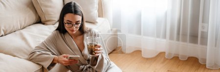 Foto de Mujer blanca bebiendo té y usando el teléfono móvil mientras está sentada en el piso en casa - Imagen libre de derechos