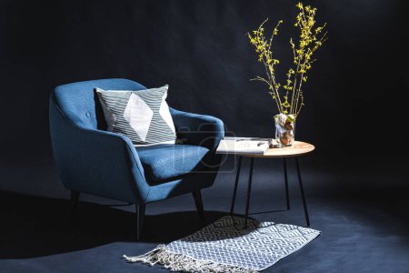 interior, vacaciones y concepto de decoración para el hogar - moderna silla azul con almohada y huevos de Pascua en jarrón con ramas forsythia y revista en la mesa en habitación oscura