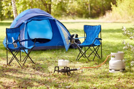 food cooking, tourism and travel concept - close up of camping pot on tourist gas burner, blue tent and two folding chairs at camp