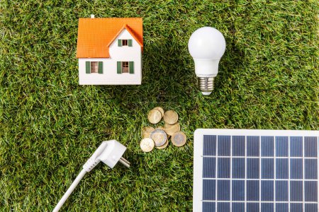 energy saving, power and sustainability concept - close up of solar battery model, lightbulb, plug with house and money on green grass background