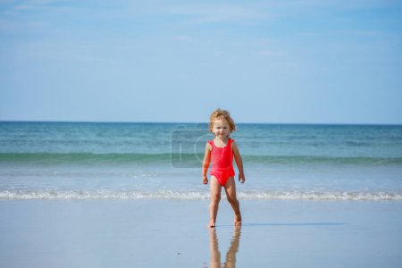 Foto de Pequeña linda chica de pie en una playa de arena sobre las olas del océano sonriendo en las vacaciones de verano - Imagen libre de derechos