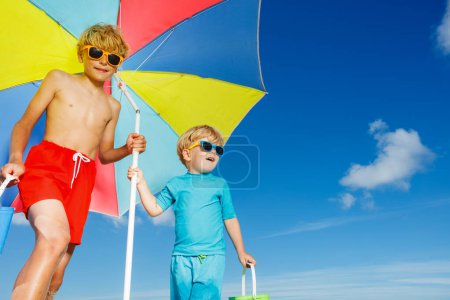 Close portrait of two boys wear sunglasses stand on the sand ocean beach under parasol