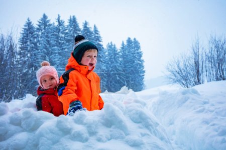 Two kids, little boy with sister play snowball sitting in the snow fortress over mountains on background